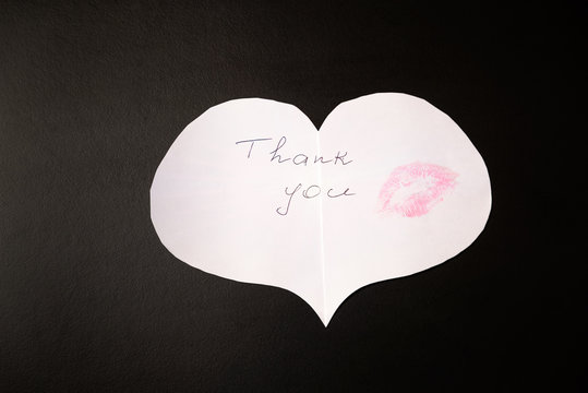 White paper origami heart with note "Thank you" and print of female lips - kiss pink color is on the black table/background. International Thank You Day	