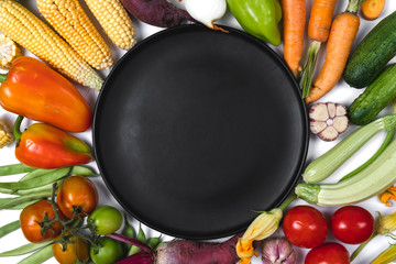 Pattern made of autumn vegetables. Tomatoes, onion, cucumber, carrot, garlic, red beet, pepper, zucchini, corn, green haricot and black plate on white background. Flat lay, top view, copy space