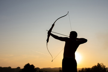Silhouette of a man with an ancient weapon bow and arrow on a background of sky and sunset - Powered by Adobe