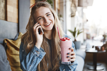 Attractive fair-haired female student in denim jacket, tilting head and smiling broadly while...