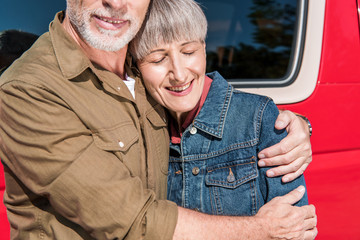 cropped view of smiling senior couple of tourists standing near red car and embracing in sunny day
