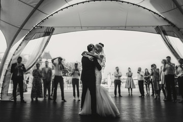 Stylish happy bride and groom gently dancing at wedding reception. Gorgeous wedding couple of newlyweds embracing while having first dance on background of guests