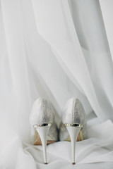 Stylish white shoes for bride on white tulle in soft morning light in hotel room. Morning preparation before wedding ceremony. Footwear for luxury event
