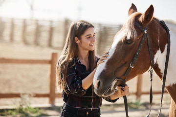 Young attractive female vet look after cute horse, petting its head, smiling tenderly. Young woman jokey became friends with pet, walking in stables, learn how to ride, hold animal on bridle - Powered by Adobe