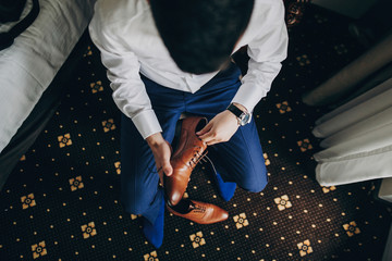 Stylish groom in blue suit putting on brown shoes near window in hotel room, top view. Morning...