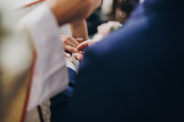Stylish wedding couple exchanging wedding rings at holy matrimony in church. Cropped view of bride and groom putting on wedding rings each other on finger. Spiritual couple