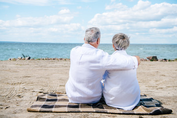 Fototapeta premium back view of senior couple in white shirts sitting on blanket and embracing at beach