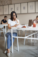 Vertical shot busy young woman in glasses sit alone cafe surrounded with working material notebook and laptop, using smartphone, scrolling social media feed during break from studying
