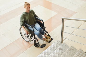 Red-haired woman sitting in wheelchair and looking at stairs she thinking how to move up the stairs at the office