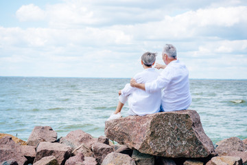 Fototapeta premium back view of senior couple in white shirts embracing near river under blue sky