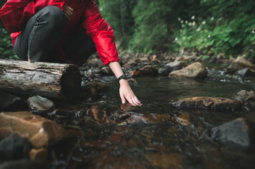 Hiker woman lowers her hand into a clear cold, mountain stream, close up photo. Hiker girl washes her hands in a mountain river. Background