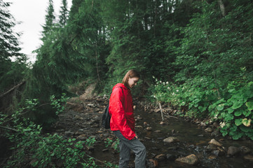 Naklejka na ściany i meble Beautiful hiker girl in a red raincoat walks in the forest in the wild,crosses a mountain stream, looks down at the water. Hiking woman in the mountains, vacation in the wild.
