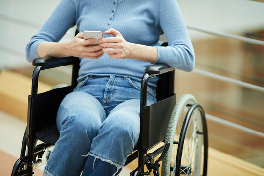 Close-up of disabled young woman in casual clothing sitting in wheelchair and communicating online on mobile phone