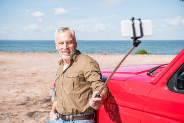 Fototapeta premium smiling senior man standing near red car and taking selfie in sunny day