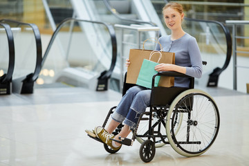 Portrait of young disabled woman sitting in wheelchair with shopping bags and smiling at camera in the modern shopping mall