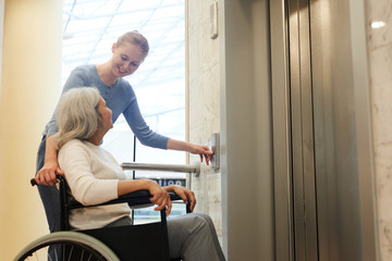 Young woman pushing the button of elevator and smiling to senior disabled woman while she sitting...