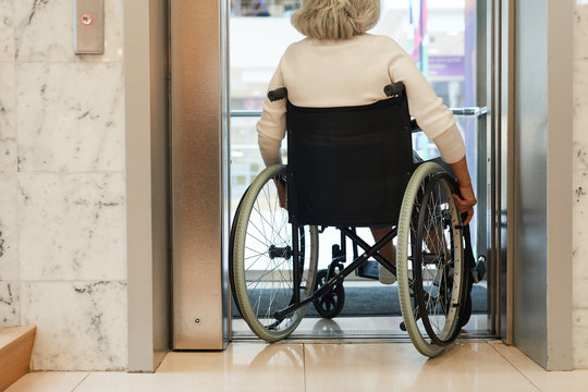 Rear View Of Senior Woman Sitting In Wheelchair And Moving Down The Elevator In Modern Building