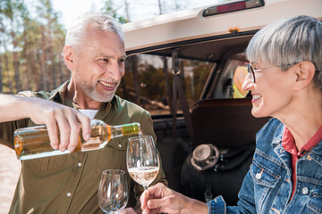 smiling senior man pouring wine in wine glasses and looking at wife near car
