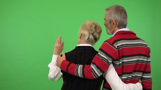 An elderly couple hugs and stands with backs to the camera and looks at the green screen background in studio - green screen studio - she points at something
