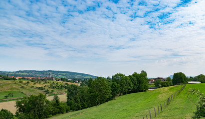 Hilly and picturesque landscape view of meadows, pastures, fields, trees, town of Brens and white clouds in the blue sky.Department of Haute-Savoie,region of Auvergne-Rhone-Alpes in France.