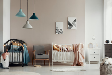 Scandi baby bedroom interior with an armchair between twin beds, standing against a beige wall with geometric paintings. Real photo