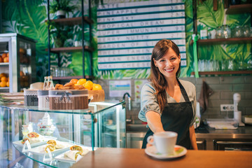 Portrait of a female barista standing behind the bar, serving coffee.