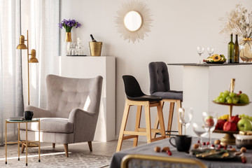 Trendy grey armchair next to two black wooden bar stools in fashionable kitchen and dining room interior