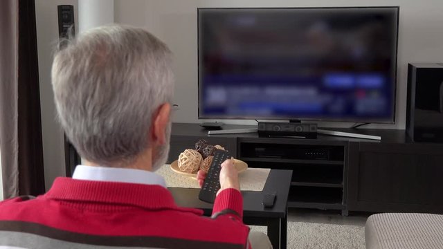 An elderly man watches out-of-focus TV and switch programs with a remote control in a living room - view from behind