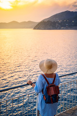 Woman in casual dress and hat with backpack looking to sea. Travel Cinque Terre in Italy in summer sunset