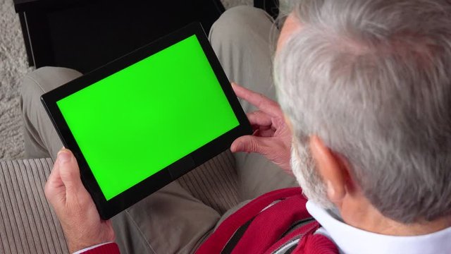 An elderly man sits on a couch in an apartment and looks at a tablet with a green screen - closeup