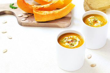 Roasted pumpkin and carrot soup with cream and pumpkin seeds on two white mug on white background with black bread slices.Vegetarian eating.Diet and healthy food concept.Space for copy and text