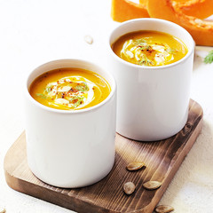 Roasted pumpkin and carrot soup with cream and pumpkin seeds on two white mug on white background with black bread slices.Vegetarian eating.Diet and healthy food concept.Space for copy and text