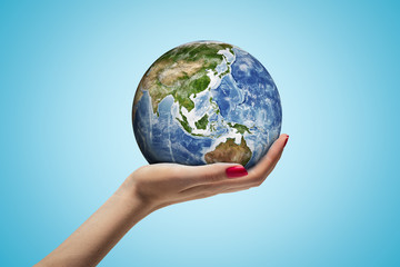 Side closeup of woman's hand facing up and holding small Earth on light blue background.