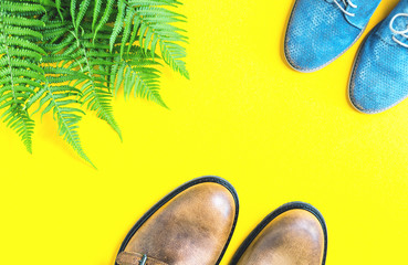 Autumn or spring trendy flat lay on bright glowing yellow. Two pairs of shoes brown male and blue female and fresh green fern leaves. copy space
