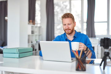 Fototapeta premium Young dedicated Caucasian male employee sitting in office, drinking coffee and using laptop. Start up business concept.