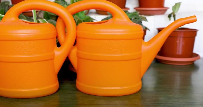 Sliding from right to left along two orange garden watering cans who are stands back to each other front of pots with flowers