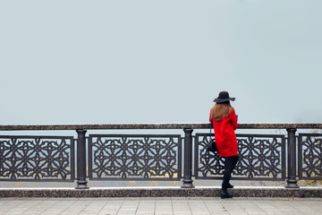 A girl in a red coat and hat, on a cold autumn morning, stands near the railing and looks into the fog. Copy space.