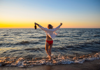 Young girl on the sea beach with a bottle of champagne and a glass, woman went into the water on the beach.