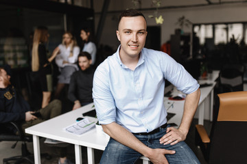 Smiling guy dressed in office style clothes is sitting on the edge of the table in the light modern office