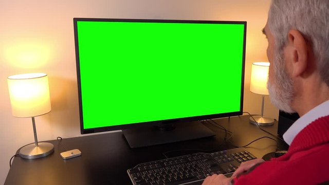 An elderly man sits in front of a computer in an apartment and looks at the green screen - closeup