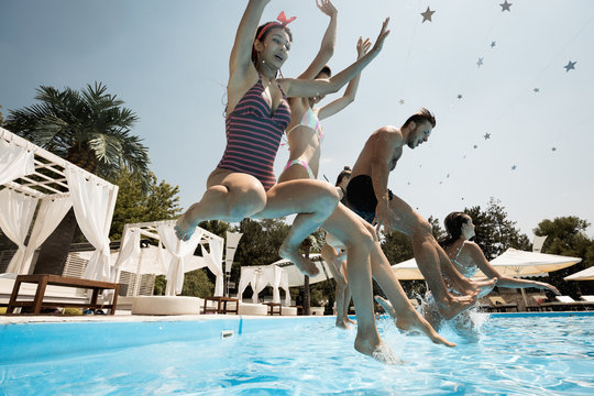 Funny company of young merry girls and guy run up to jump into the swimming pool on the open air on a sunny summer day next to the lounge zone