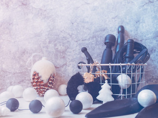 Various sex toys (handcuffs, dildo, vibrator) are in a white metal basket. Nearby is a large fir cone and Christmas balls. Gray background with cracks and scuffs. Minimal composition.
