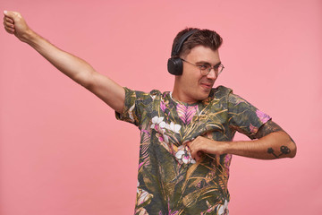 Short-haired hansome guy posing over pink background in fowered t-shirt, dancing and enjoying the music in headphones, looking away, being in nice mood