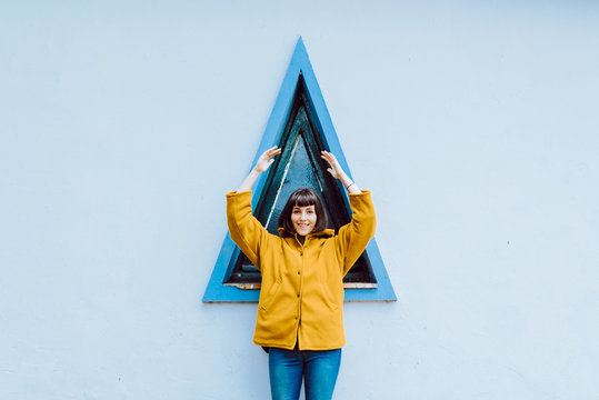 Young woman in yellow warm coat smiling and looking at camera while standing against triangle window and gray wall of building