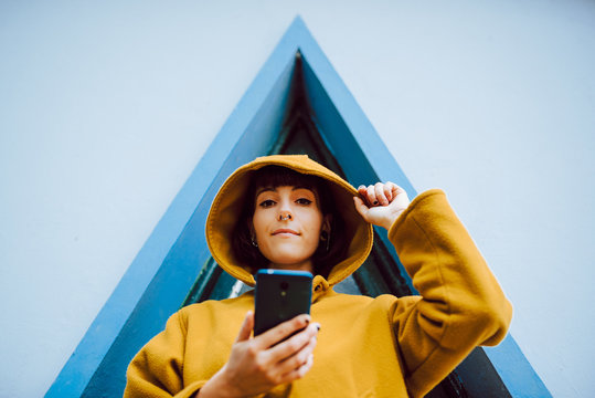 From below young female adjusting hood of yellow warm coat and looking at camera while browsing smartphone near building with triangle window