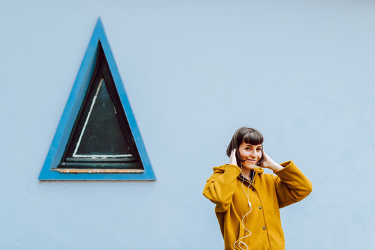 Cheerful female in yellow warm jacket smiling and touching hair while listening to music near gray building with triangle window
