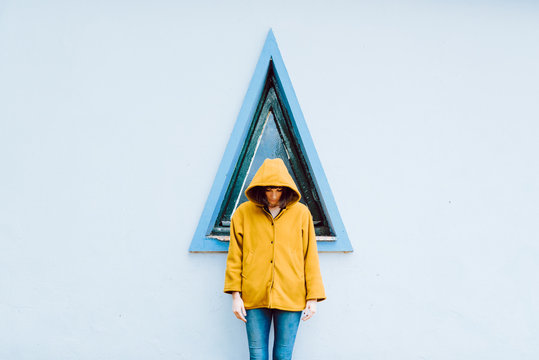 Young woman in yellow warm coat smiling and looking down while standing against triangle window and gray wall of building