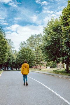 Back view of female in yellow coat walking along asphalt road on green suburban street
