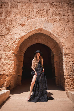 Barefoot beautiful woman in black dress and hat posing by gate of old castle on sunny day