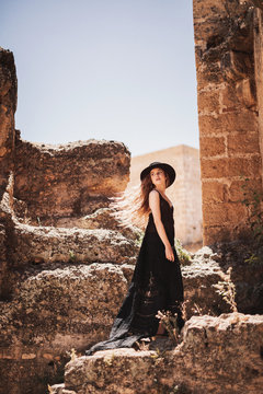 Side view of stylish woman in long black dress and hat standing by ruined building and looking back